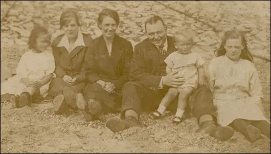 Muriel Hughes (sitting on the far left) posing on a beach with her mother and father, Elizabeth and Thomas Hughes (sitting in the middle of the picture) in a photograph taken in 1922. Tom Hughes, Muriel's father, holds his son, two-year-old John Hughes. The girl sitting at the extreme right of the picture is Muriel's cousin Charlotte. Another relative, Violet, sits between Muriel and her mother.